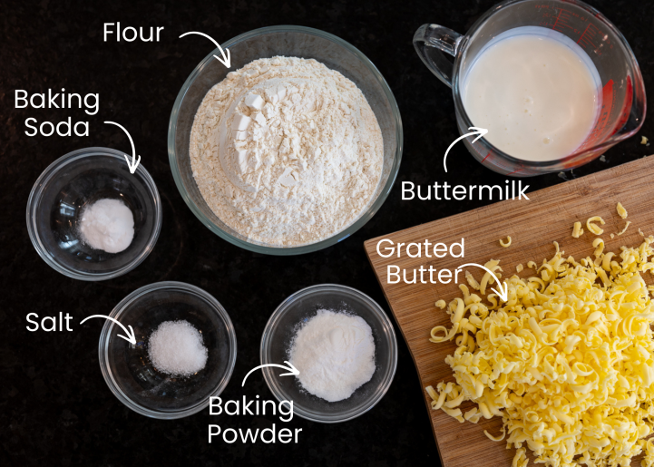 A photo with ingredients in clear bowls on a black surface. Each ingredient is labeled. The ingredients are for southern buttermilk biscuits.