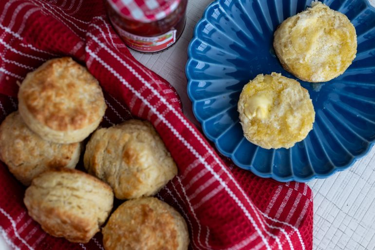 A blue plate with a split biscuit with butter, and a basket with a red cloth with whole biscuits. This is a recipe for southern buttermilk biscuits.