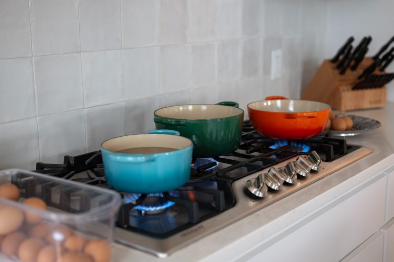 A photo of a kitchen cooktop with three different colored pots on it. There is a flame under each of the pots and you can see eggs to the side. This is on a post about testing 3 hardboiled egg methods. How to boil eggs so that the shells peel off easily.