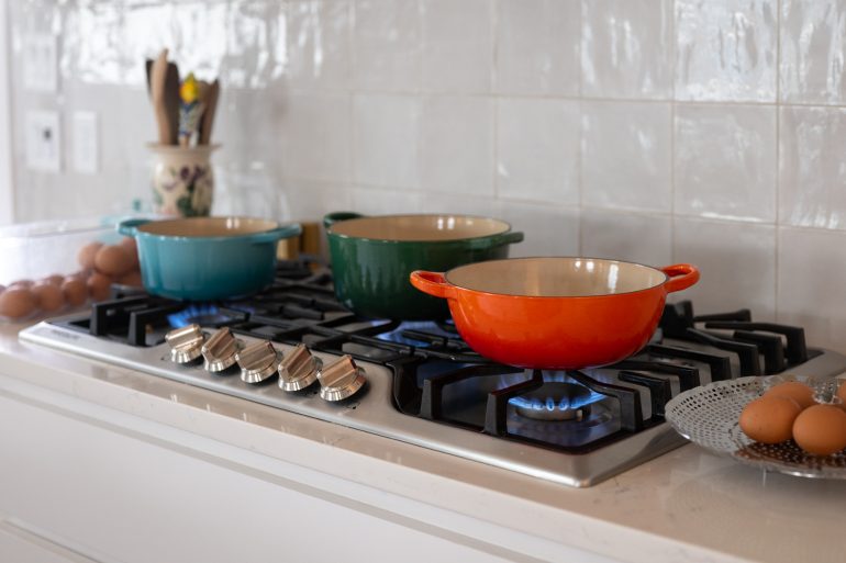 A photo of a kitchen cooktop with three different colored pots on it. There is a flame under each of the pots and you can see eggs to the side. This is on a post about testing 3 hardboiled egg methods. How to boil eggs so that the shells peel off easily.