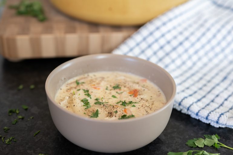 Photo of a bowl of chicken and dumplings. You can see the pot in the background and there is a blue and white tea towel to the side. 

