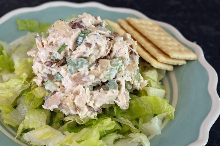 A blue plate with white trim on a black background. The plate has a bed of lettuce with classic chicken salad on it.