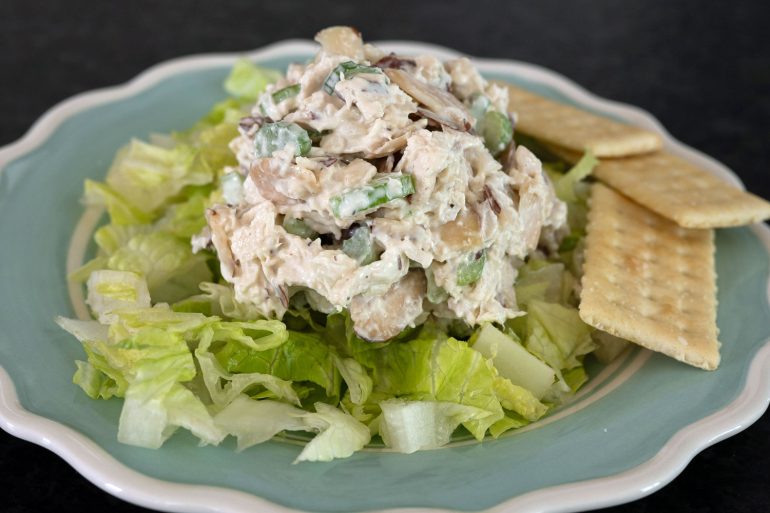 A blue plate with white trim on a black background. The plate has a bed of lettuce with classic chicken salad on it and some club crackers on the side. The chicken salad has chicken, celery, and almonds.