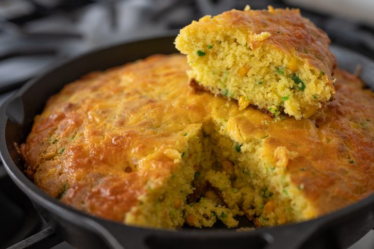 Cornbread in an iron skillet. There is a wedge of cornbread cut out and sitting on top. You can see kernels of corn and diced jalapeno in the cornbread.