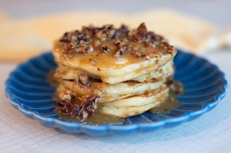 A blue plate with a stack of 4 fluffy pancakes topped with a sauce with pecans.