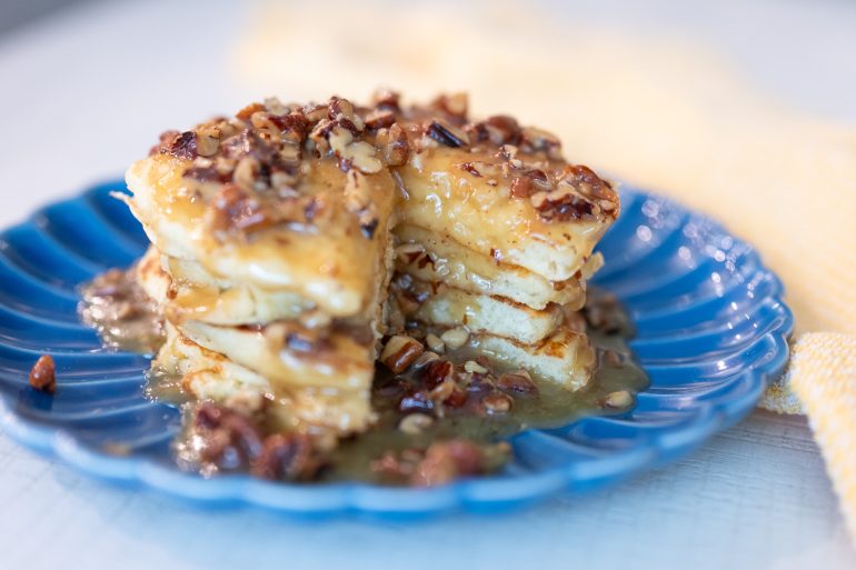 A stack of pancakes with a wedge cut out that show the fluffy interior of the pancakes. There is a sauce over the pancakes with pecans.