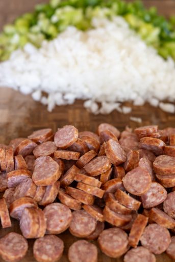 A cutting board that shows sliced sausage with chopped onion, celery, and green pepper in the background.