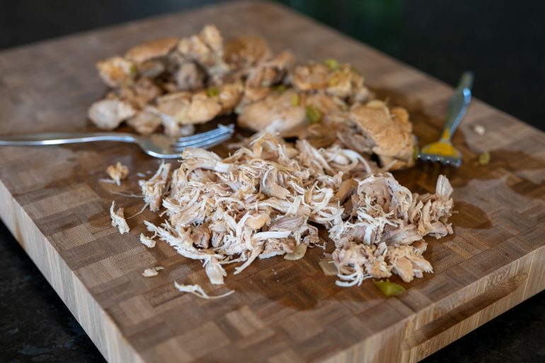 A cutting board with cooked chicken on it. The chicken is partially shredded and there are also two forks on the cutting board.