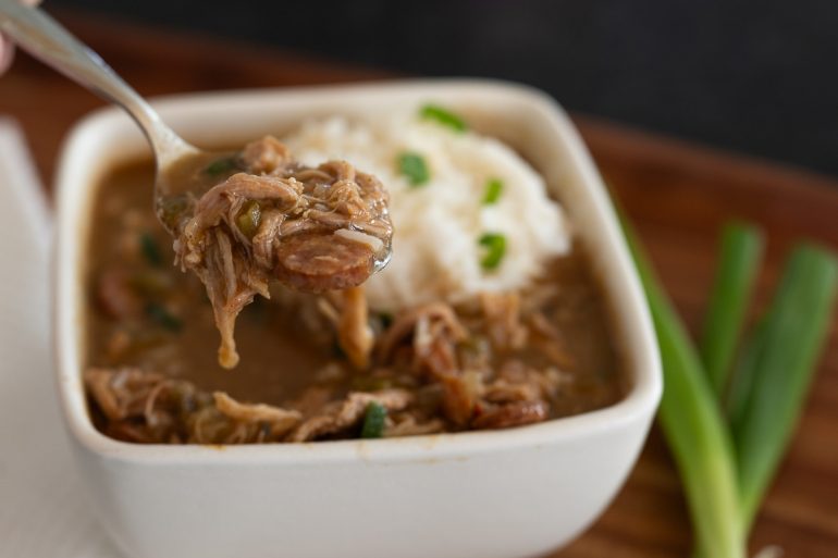 A square bowl of chicken and sausage gumbo. A spoonful of gumbo is clear in the foreground lifted out of the bowl with the rice and remaining gumbo blurred in the background.