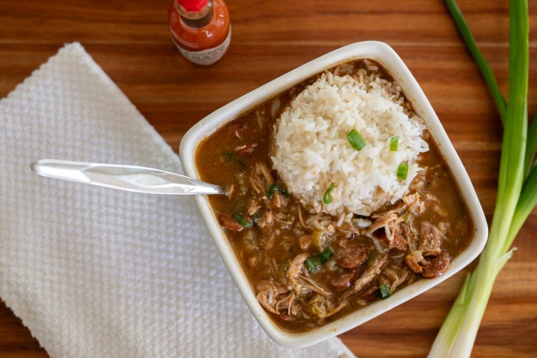 Photo shot from above. There is a square bowl of chicken and sausage gumbo with rice on the right side and a spoon on the left. You can also see a green onion on the far right and a white tea towel on the left with a bottle of hot sauce on the upper left just barely showing.