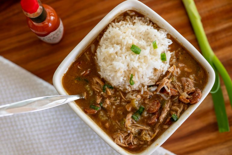 Photo shot from above. There is a square bowl of chicken and sausage gumbo with rice on the right side and a spoon on the left. You can also see a green onion no the far right and a white tea towel on the left with a bottle of hot sauce on the upper left just barely showing.