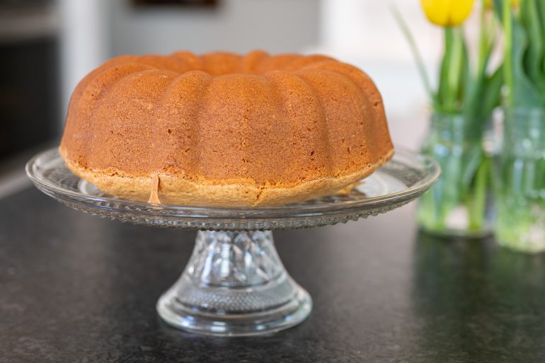A classic cream cheese pound cake that has been cooked in a bundt pan. It is sitting on an elevated glass cake plate on a counter.