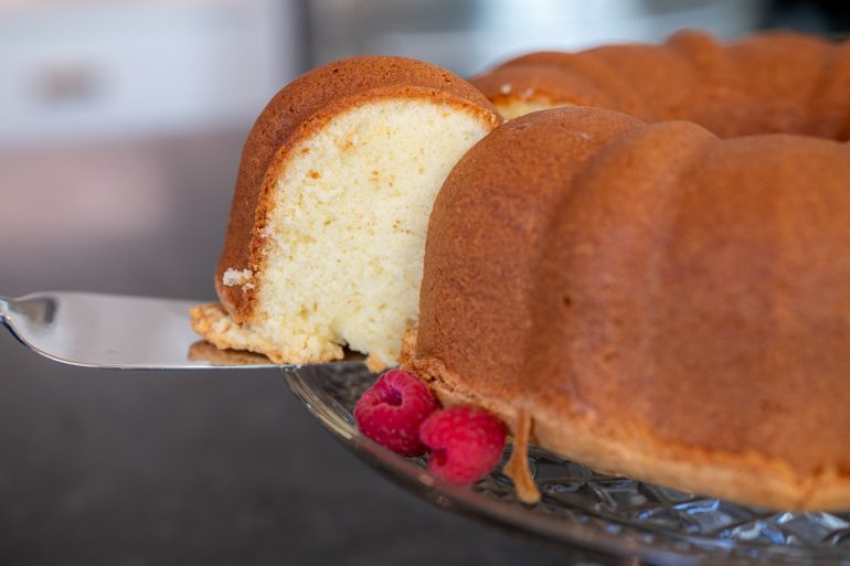 A classic cream cheese pound cake sitting on a crystal cake plate that has been shot from the side. A piece of the cake has been cut and there is a cake server removing the piece. You can see the piece of the cake from the side and it is about halfway removed.
