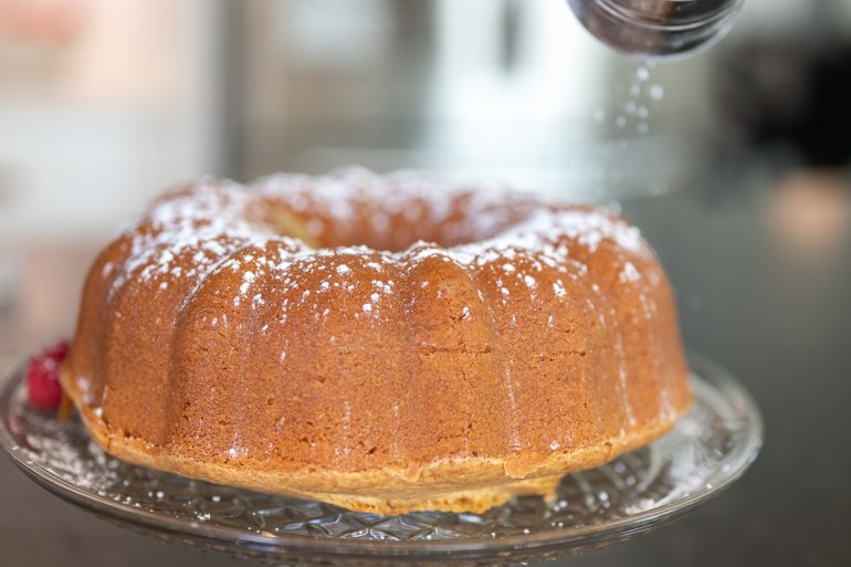 A close up of a cake that has been cooked in a bundt pan. In the upper right corner is a shaker that is shaking powdered sugar onto the cake.