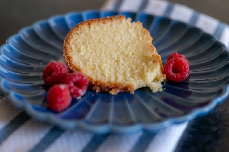 A classic cream cheese pound cake on a blue plate with raspberries on the sides. The cake is dense but moist and is shot from slightly above.