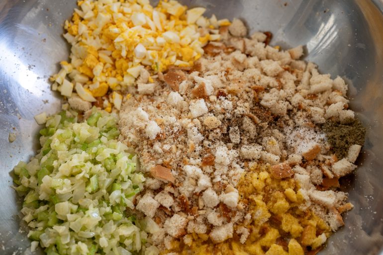 A big silver bowl with sautéed onion and celery, chopped boiled egg, some spices, cornbread and white bread, and a raw egg. It looks like the mixture will soon be mixed up.