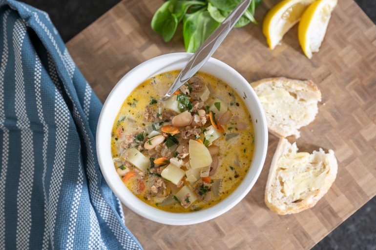 Italian sausage, white bean, and potato soup. The photo is of a bowl of soup from above. It looks rich and delicious. You see some torn bread on the right side. A blue tea towel on the left and some lemon in the upper right corner.