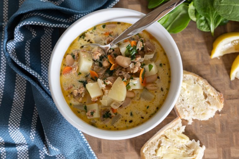 Italian sausage, white bean, and potato soup. The photo is of a bowl of soup from above. It looks rich and delicious. You see some torn bread on the right side. A blue tea towel on the left and some lemon in the upper right corner.