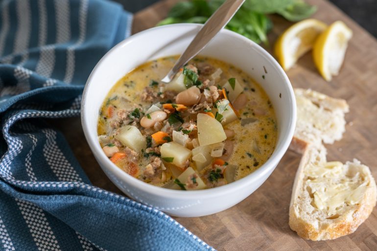 Italian sausage, white bean, and potato soup. The photo is of a bowl of soup from above. It looks rich and delicious. You see some torn bread on the right side. A blue tea towel on the left and some lemon in the upper right corner.