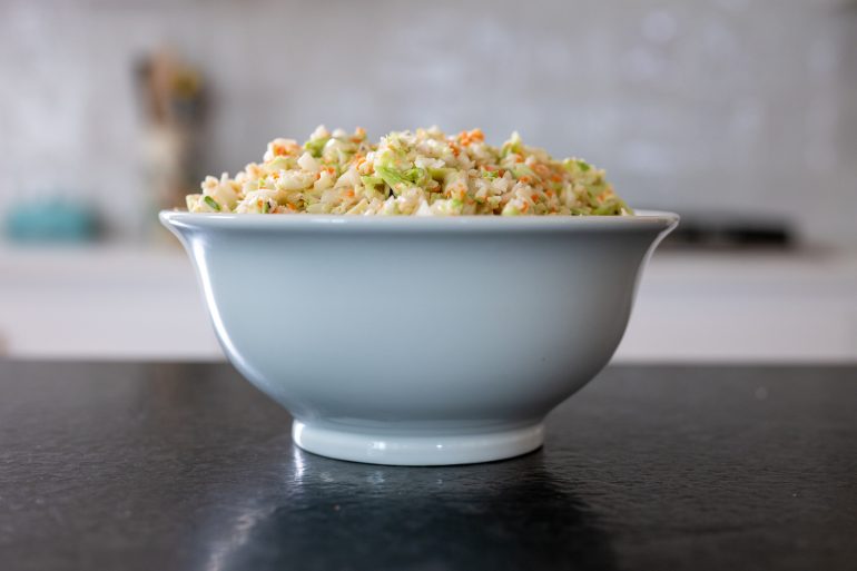 This KFC style Coleslaw is shown in a white bowl. You are seeing it from the side. The background is blurry.