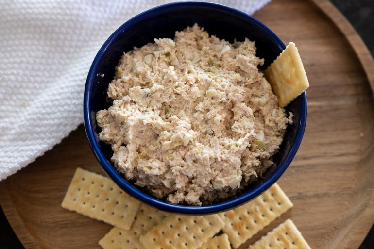 A blue bowl of smoked turkey dip on a wooden charger with a tea towel in the upper left corner. Club Crackers in the bottom of the photo and one cracker in the dip.