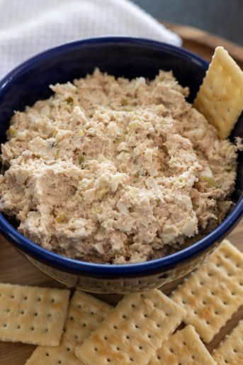 A photo of smoked turkey dip in a blue bowl. It is on a wooden charger with a white tea towel in the upper left corner. A cracker in the dip and several crackers in front of the bowl.