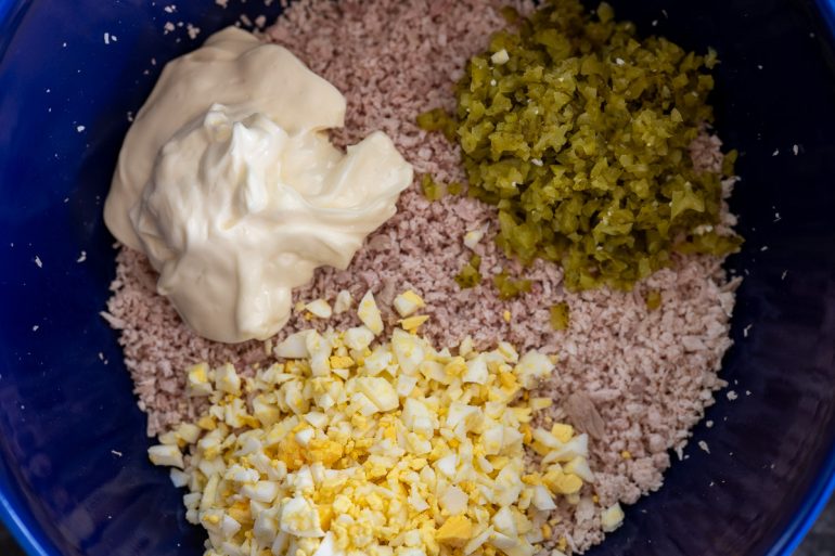A shot taken from above of a bowl of finely chopped turkey. You can see mayonnaise, chopped boiled eggs, and chopped pickle which will be stirred into the turkey.