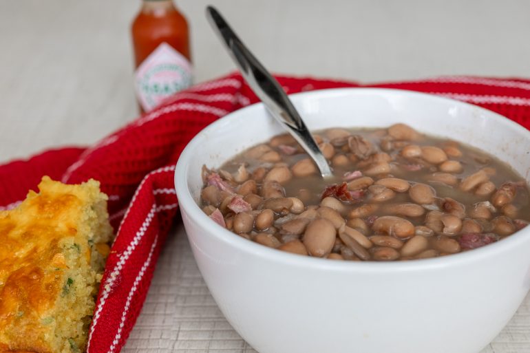 Pinto beans in a white bowl. There is a red tea towel in the background and you can see tabasco sauce blurred at the back with a piece of cornbread on the lower left corner.