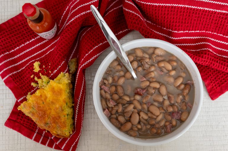 Overhead shot of southern pinto beans in a white bowl. There is cornbread on the left side of the bowl on a red tea towel. 