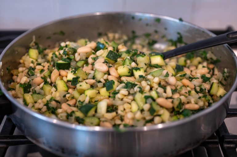 A mixture in a skillet of white beans, zucchini, onion, corn, and spinach.