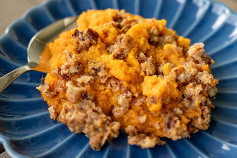 A top down view of a blue plate with a vibrant orange sweet potato casserole on it. There is a spoon on the side of the plate and the sweet potatoes have been topped with pecans.
