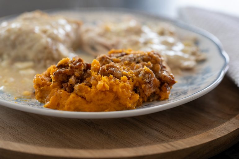 A side view of a plate. There is a sweet potato casserole with pecan topping closest to the camera. In the background you can see dressing and turkey covered in gravy.