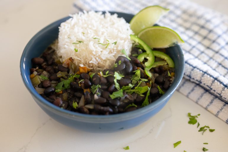 Cuban black bean soup with rice in a blue bowl. The beans are sprinkled with cilantro and a few jalapeños with some limes on the side. 