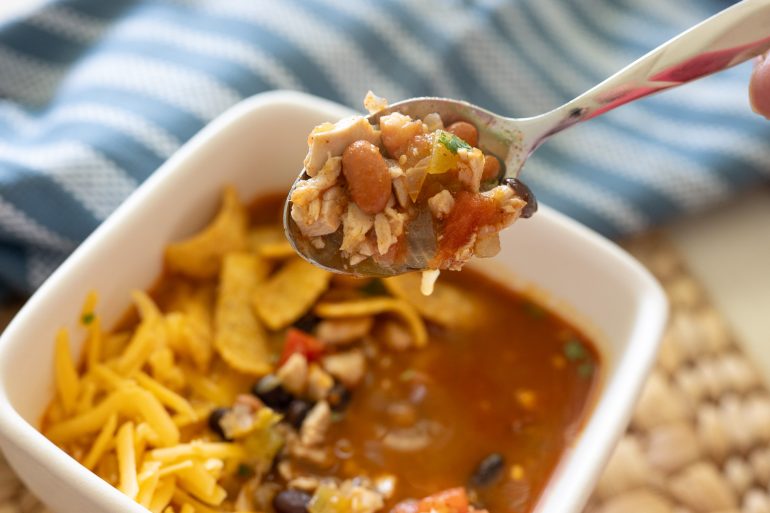 A white filled with Chicken taco soup. It is sitting on a rustic placemat with a blue tea towel in the background. There is a spoonful of soup lifted toward the reader. Fritos and some shredded cheddar are down in the bowl. On the spoon you can see chicken, black bean, pinto beans, and tomato.
