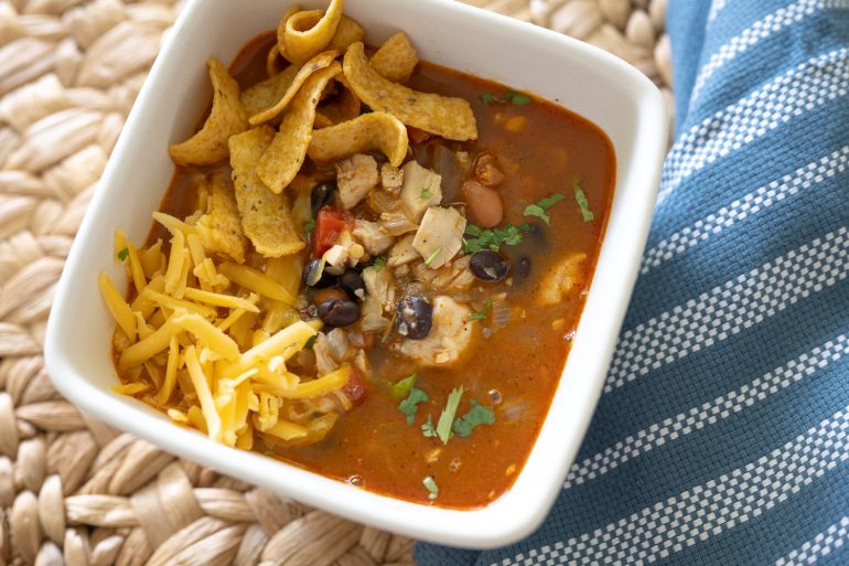 A white filled with soup. It is sitting on a rustic placemat with a blue tea towel in the background. The soup has cilantro sprinkled on top, Fritos and some shredded cheddar. You can see chicken, black bean, pinto beans, and tomato.