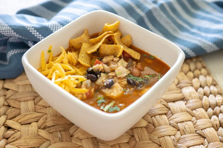 A white bowl of easy chicken taco soup. The soup has cilantro sprinkled on top, Fritos in the back. And some shredded cheddar. You can see chicken, black bean, pinto beans, and tomato. It looks frothy and flavorful. 