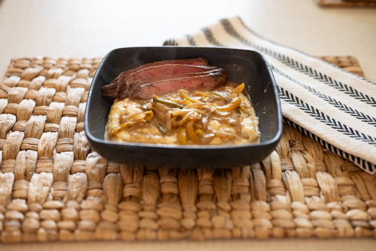 A side view of a black bowl filled with cheddar cheese grits which are topped with a pepper and onion sauce and medium rare flank steak. There is a black and white tea towel on the right side. This photo is on a recipe for creole flank steak over cheddar grits with creamy pan sauce. 