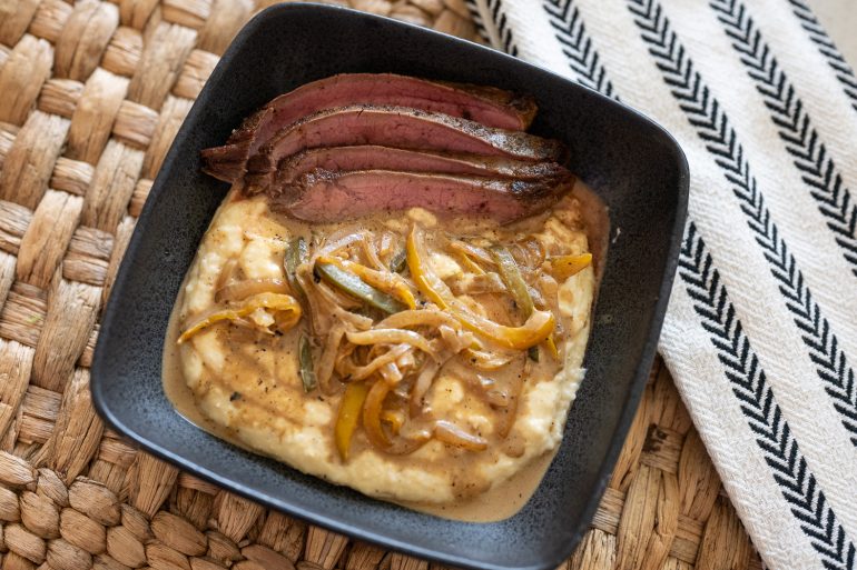 bowl of cheddar grits topped with onion pepper sauce and flank steak. 