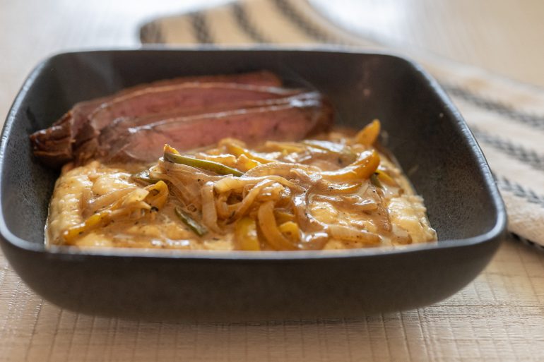 a closeup side view of a black bowl filled with cheddar cheese grits which are topped with a pepper and onion sauce and medium rare flank steak. There is a black and white tea towel on the right side. This photo is on a recipe for creole flank steak over cheddar grits with creamy pan sauce. 
