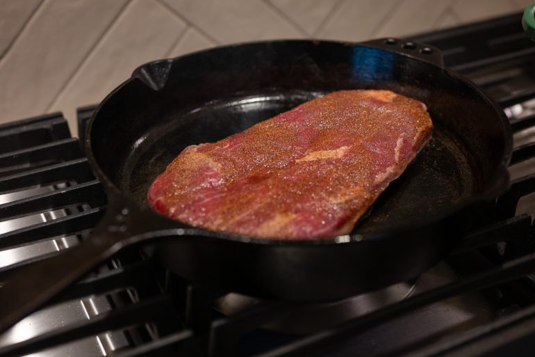 A flank steak in a cast iron skillet on a stove top.