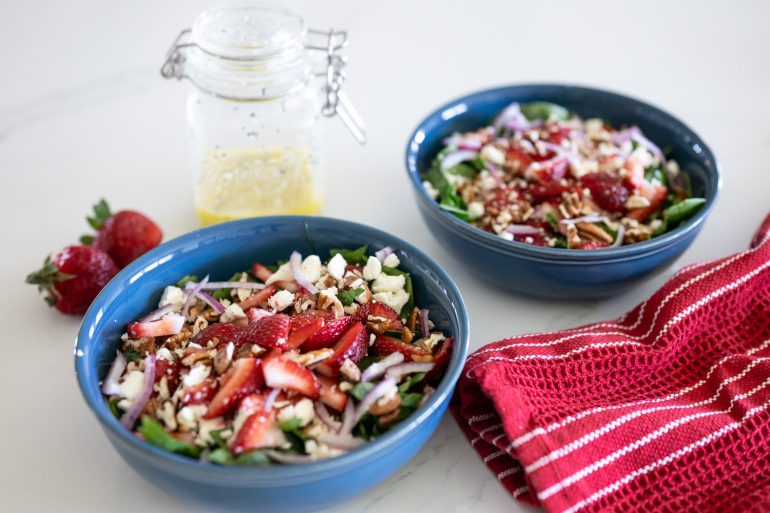 2 bowls filled with salad. You can see spinach, strawberries, feta cheese, pecans, and thinly sliced red onion. There is a mason jar of dressing in the background. This is strawberry spinach salad. 
