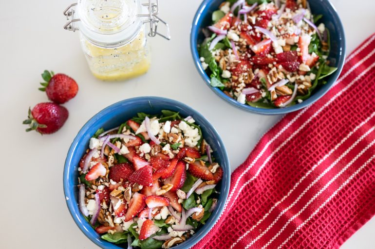 2 blue bowls filled with a salad that contains spinach, strawberries, red onion, pecans, and feta cheese. There is a red tea towel in the bottom right corner and you can see a mason jar with white wine poppy seed vinaigrette. 