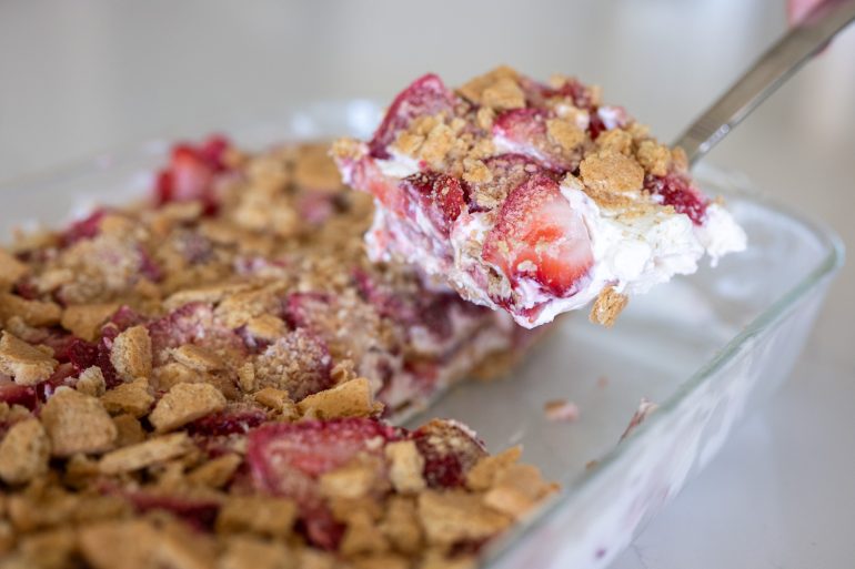 la piece of strawberry ice lifted out of a glass baking dish on a spatula. with strawberries, whipping cream, and graham crackers. 