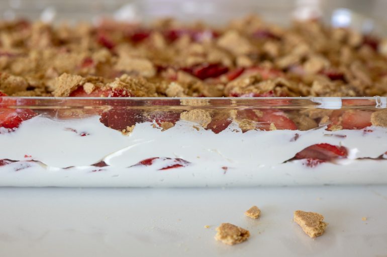 A layered dessert in a glass baking dish. You can see a few pieces of graham cracker on the counter in the foreground then in the background is the dish with whipped cream, strawberries and graham crackers. 
